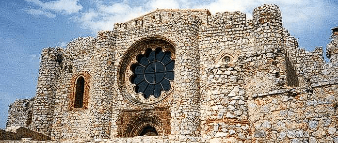 Primer plano de la fachada de piedra del Sacro Convento-Castillo de Calatrava la Nueva. Destaca un gran rosetón central de estilo gótico con tracería lobulada, flanqueado por muros robustos de piedra irregular y una ventana de arco apuntado a la izquierda bajo un cielo azul con nubes.