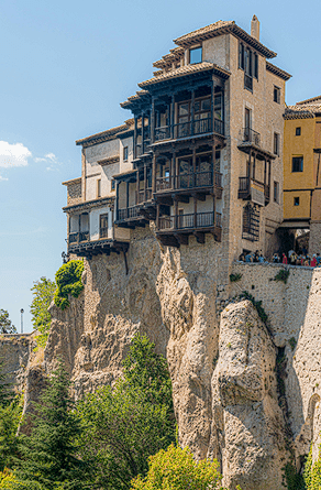 Fotografía vertical de las Casas Colgadas de Cuenca. Se ven varios edificios de piedra y mampostería con balcones de madera oscura que sobresalen directamente sobre un alto acantilado rocoso. En la base del desfiladero hay vegetación verde y árboles, bajo un cielo azul despejado.