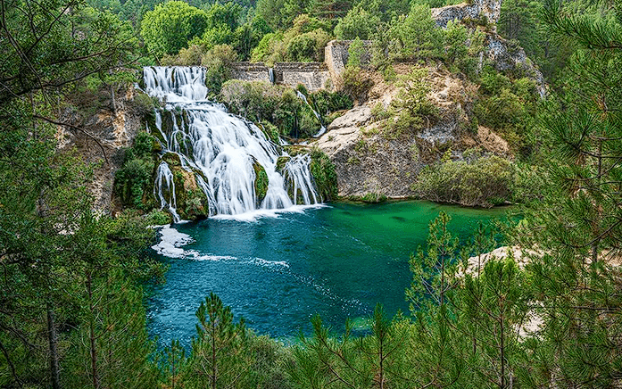 Paisaje natural que muestra una cascada de varios niveles cayendo sobre una poza de aguas cristalinas de color turquesa. La cascada está rodeada de exuberante vegetación verde y formaciones rocosas. En el primer plano, unas ramas de pino enmarcan la vista, y al fondo se aprecia un denso bosque de pinos bajo una luz diurna suave.
