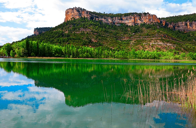 Paisaje panorámico de la Laguna de Uña. En el primer plano, aguas tranquilas de color verde esmeralda reflejan el cielo azul y las montañas. A la derecha, asoman unas cañas secas desde el agua. Al fondo, una hilera de chopos verdes precede a una imponente montaña con paredes de roca caliza naranja y cima plana, característica de los relieves de la zona.