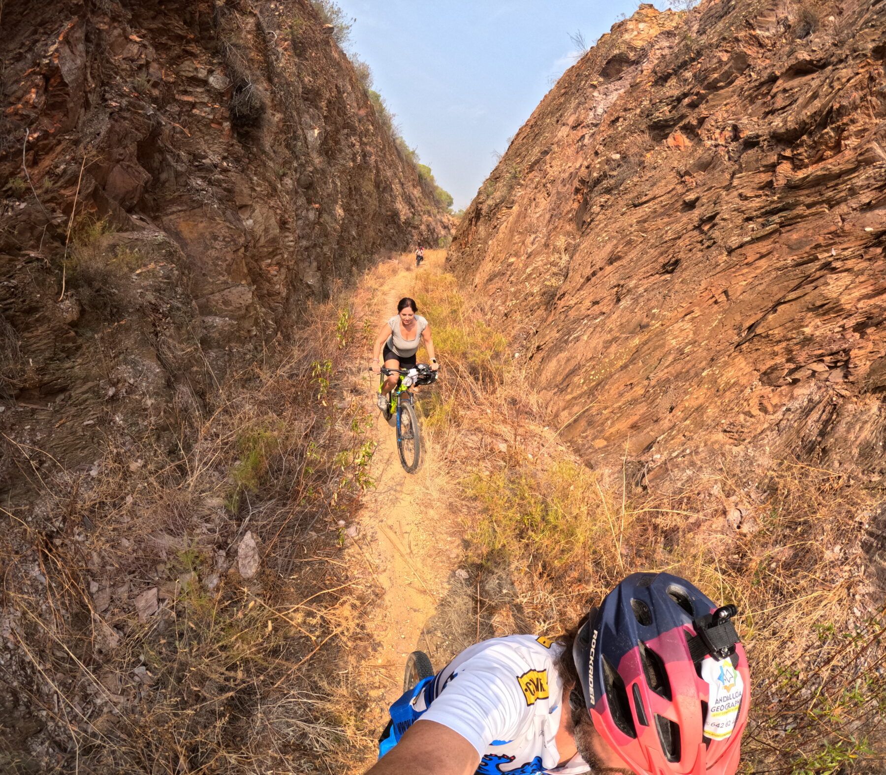 Tomada con GoPro desde el interior de un corredor natural excavado en la roca, una ciclista desciende un sendero estrecho y empinado rodeado de paredes de piedra roja. Una imagen de pura adrenalina en la Vía Verde.