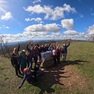 El grupo celebra la llegada a la cima del Bonales levantando los brazos y ondeando la bandera de Andalucia Geographic, con un amplio panorama sobre las colinas de la Sierra de Aracena al fondo.