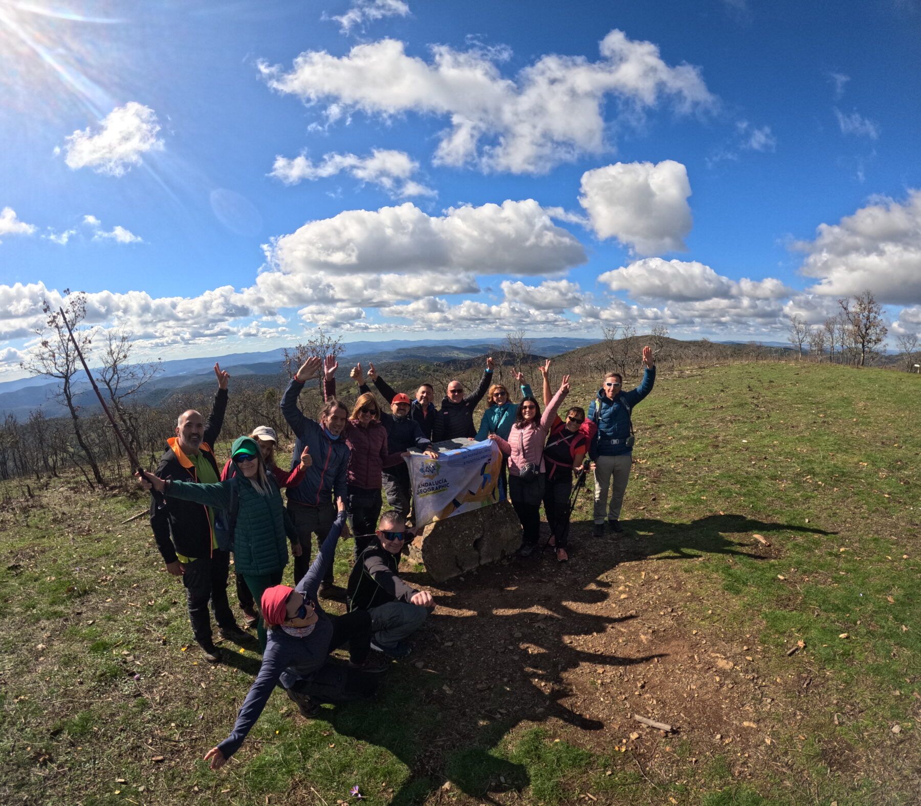 El grupo celebra la llegada a la cima del Bonales levantando los brazos y ondeando la bandera de Andalucia Geographic, con un amplio panorama sobre las colinas de la Sierra de Aracena al fondo.