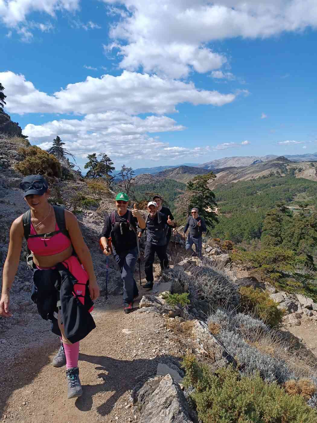 Grupo de senderistas en ruta: Un grupo de cinco personas avanza en fila por un sendero de tierra y piedra que bordea la ladera de una montaña. Se muestran sonrientes y con una actitud alegre, con algunos de ellos haciendo el gesto de "pulgar arriba" hacia la cámara. Primer plano: Una mujer lidera el grupo vistiendo un top deportivo de color rosa vibrante, mallas a juego, una gorra negra con gafas de sol encima y una chaqueta oscura atada a la cintura. Acompañantes: Detrás de ella, cuatro hombres caminan por el sendero estrecho, equipados con ropa técnica de montaña, mochilas de senderismo y bastones de trekking. Paisaje de la Sierra de las Nieves: El entorno muestra una vegetación típica de alta montaña con matorrales bajos, rocas calizas y algunos pinsapos dispersos en la pendiente. Fondo panorámico: Al fondo se aprecia un vasto valle verde rodeado de montañas que se extienden hacia el horizonte, bajo un cielo azul brillante con nubes blancas algodonosas. Condiciones climáticas: La iluminación clara y las sombras definidas indican un día soleado y despejado, ideal para la práctica del senderismo en la zona del Torrecilla.