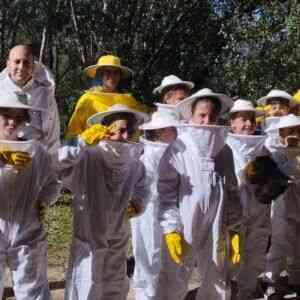 Un grupo de una decena de participantes, adultos y niños mezclados, posa orgullosamente ante la cámara en un entorno natural boscoso y soleado. Todos están equipados con trajes apícolas completos, algunos en blanco y otros en amarillo vivo, con sombreros de velo protector y guantes a juego. Sus gestos expresivos y su visible entusiasmo dan testimonio de una experiencia pedagógica lúdica e inolvidable en torno al fascinante mundo de las abejas, en el marco del programa de educación ambiental para escolares propuesto por Andalucia Geographic.