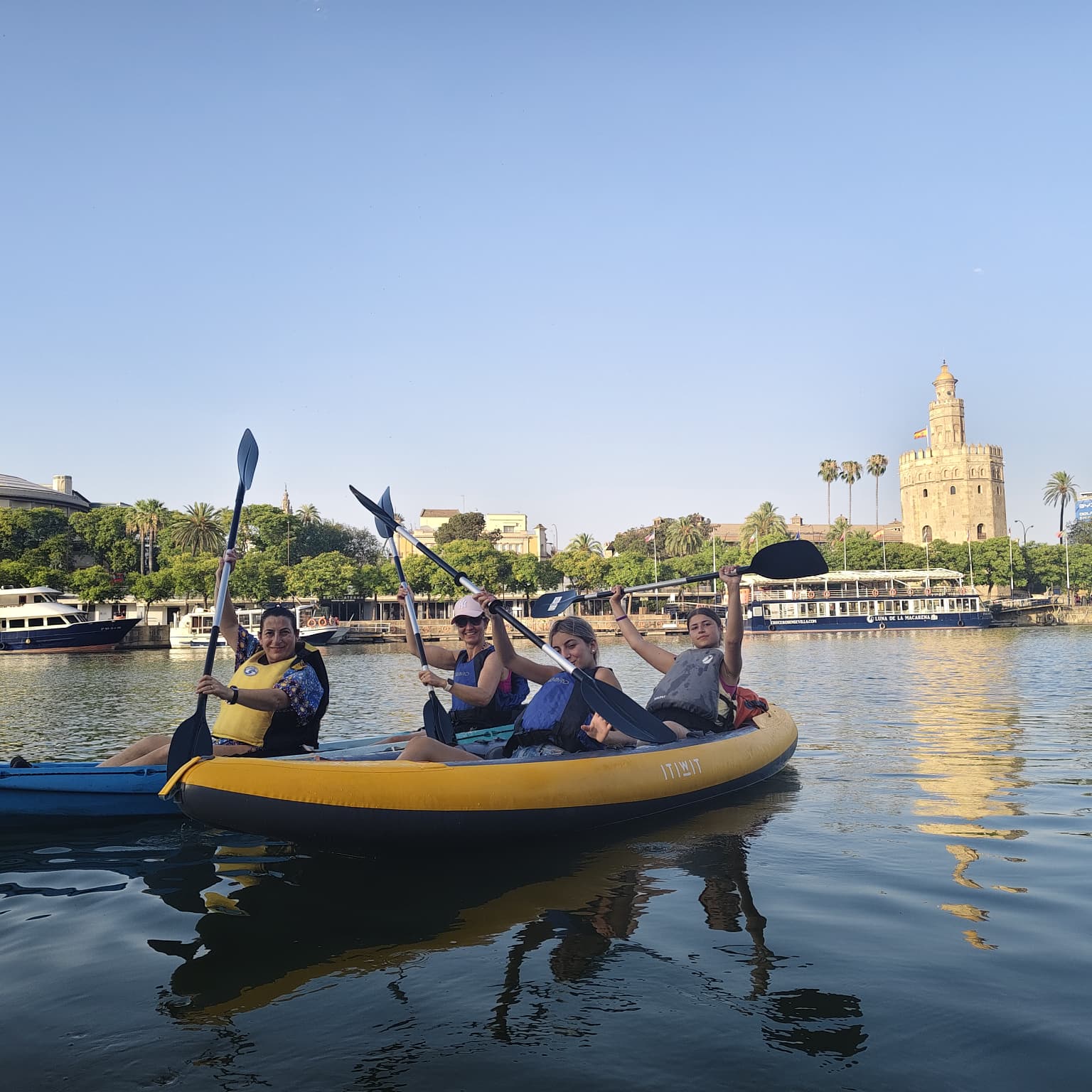 Cuatro participantes a bordo de un gran kayak hinchable levantan sus remos con entusiasmo en el Guadalquivir, con la Torre del Oro reflejándose en el agua.