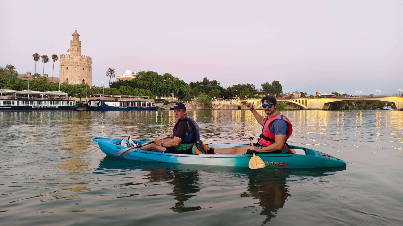 Dos participantes reman en kayak por el Guadalquivir con la célebre Torre del Oro iluminada al atardecer al fondo. Un momento mágico en el corazón de Sevilla.