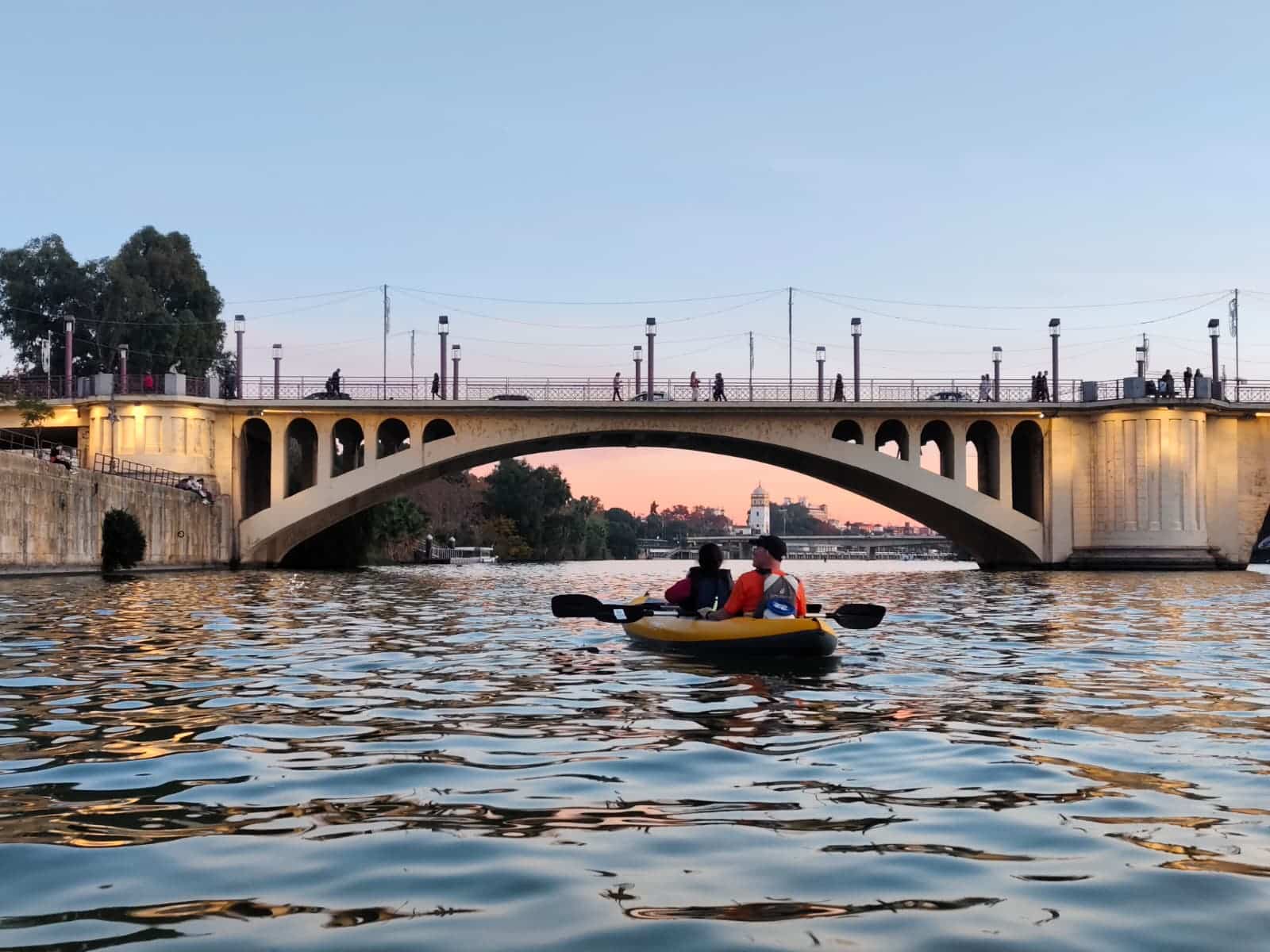 Dos participantes cruzan en kayak bajo un gran puente de arco de Sevilla, bañado en los tonos anaranjados del atardecer. Una imagen poética y luminosa.