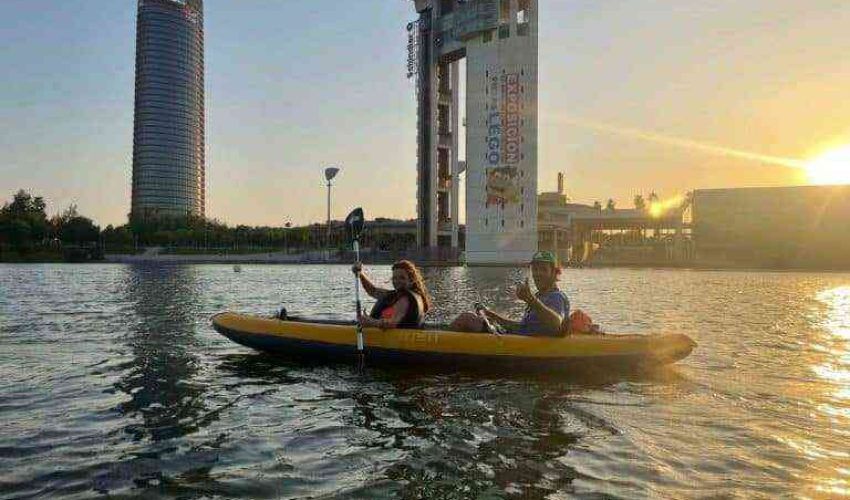 Dos kayakistas, radiantes de felicidad, reman codo a codo por el Guadalquivir en dirección a la espectacular puesta de sol que enciende el horizonte. La Torre Sevilla, reconocible edificio de gran altura de la capital andaluza, y estructuras modernas se recortan a contraluz en esta intensa y dorada luz. Uno de los kayakistas levanta su pala en señal de alegría, expresando a la perfección el entusiasmo que procura esta única actividad náutica en el corazón de la ciudad histórica de Sevilla.
