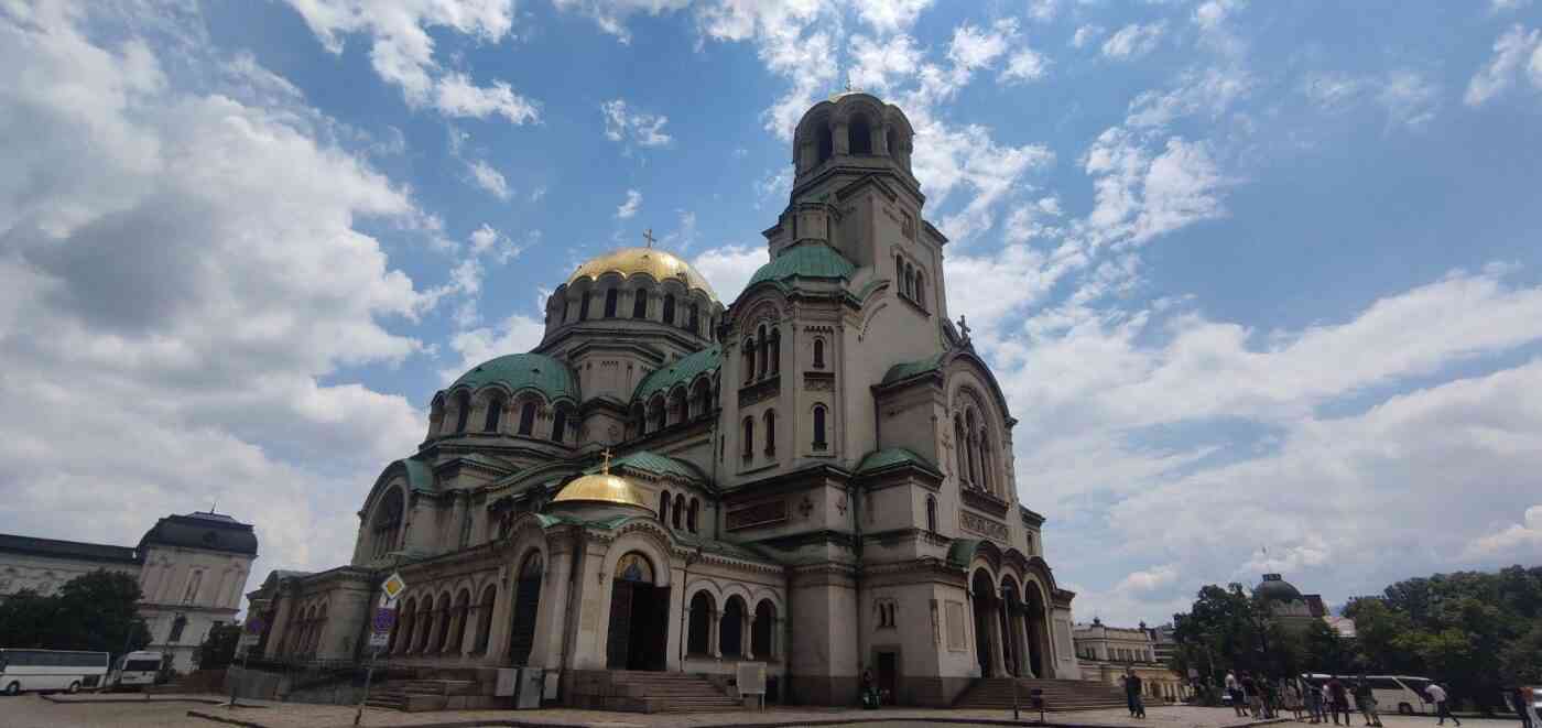 Fachada y cúpulas doradas y verdes de la Catedral de San Alejandro Nevski en Sofía bajo un cielo azul con nubes.
