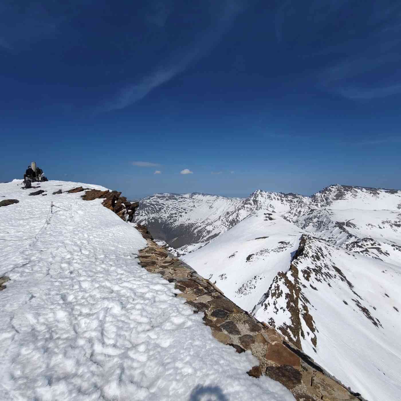 Una impresionante vista panorámica desde la cima de una montaña nevada en Sierra Nevada, España. En primer plano, una cresta rocosa y afilada cubierta parcialmente por nieve se extiende hacia el centro de la imagen, creando un fuerte contraste entre la roca oscura y el blanco brillante. Al fondo, se observan imponentes picos montañosos, como el Mulhacén y la Veleta, bajo un cielo azul despejado y profundo. El paisaje transmite una sensación de altitud, serenidad y la magnitud de la alta montaña durante el invierno o la primavera temprana.
