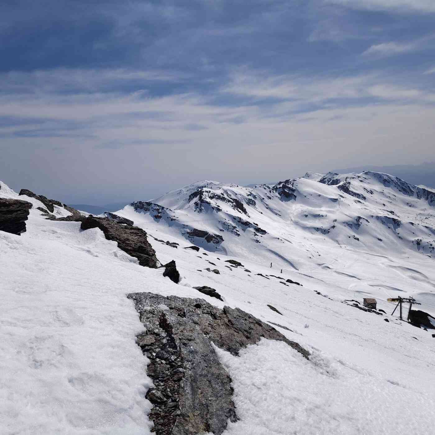 Una impresionante vista panorámica desde la cima de una montaña nevada en Sierra Nevada, España. En primer plano, una cresta rocosa y afilada cubierta parcialmente por nieve se extiende hacia el centro de la imagen, creando un fuerte contraste entre la roca oscura y el blanco brillante. Al fondo, se observan imponentes picos montañosos, como el Mulhacén y la Veleta, bajo un cielo azul despejado y profundo. El paisaje transmite una sensación de altitud, serenidad y la magnitud de la alta montaña durante el invierno o la primavera temprana.