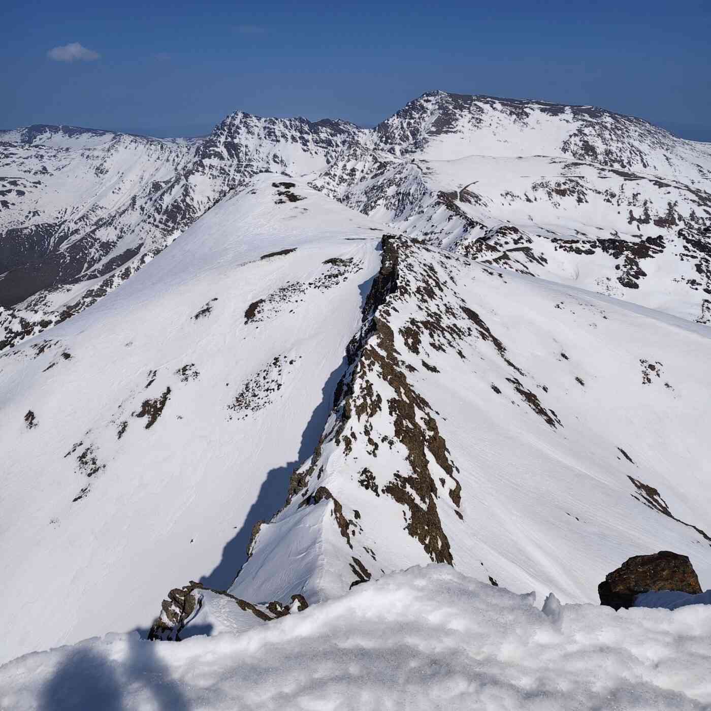 Una impresionante vista panorámica desde la cima de una montaña nevada en Sierra Nevada, España. En primer plano, una cresta rocosa y afilada cubierta parcialmente por nieve se extiende hacia el centro de la imagen, creando un fuerte contraste entre la roca oscura y el blanco brillante. Al fondo, se observan imponentes picos montañosos, como el Mulhacén y la Veleta, bajo un cielo azul despejado y profundo. El paisaje transmite una sensación de altitud, serenidad y la magnitud de la alta montaña durante el invierno o la primavera temprana.